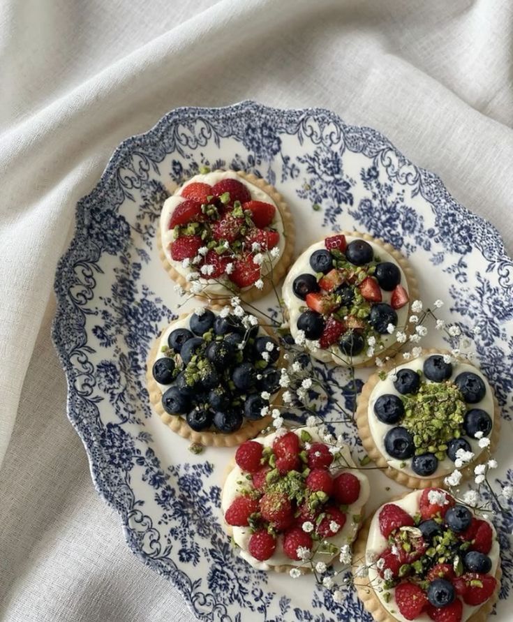 A decorative plate with assorted tartlets topped with fresh berries and pistachios, garnished with tiny white flowers, set on a light background.