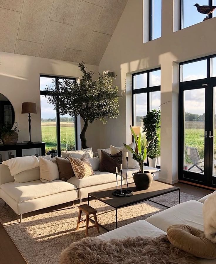 A cozy living room featuring neutral-colored sofas adorned with various cushions, a wooden coffee table, and plants, illuminated by large windows, allowing natural light to flow in.
