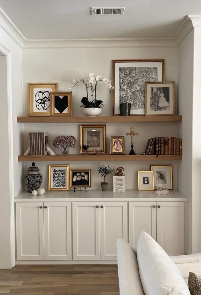 A stylish interior display featuring a wooden shelf with framed art, a potted orchid, decorative objects, and books arranged neatly above white cabinetry.