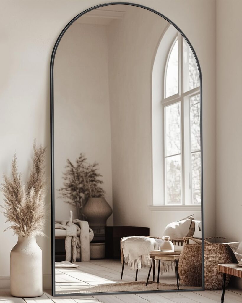 A large arched mirror reflects a well-decorated room featuring neutral tones, natural textures, and cozy furnishings, including a vase with pampas grass and a stylish chair.