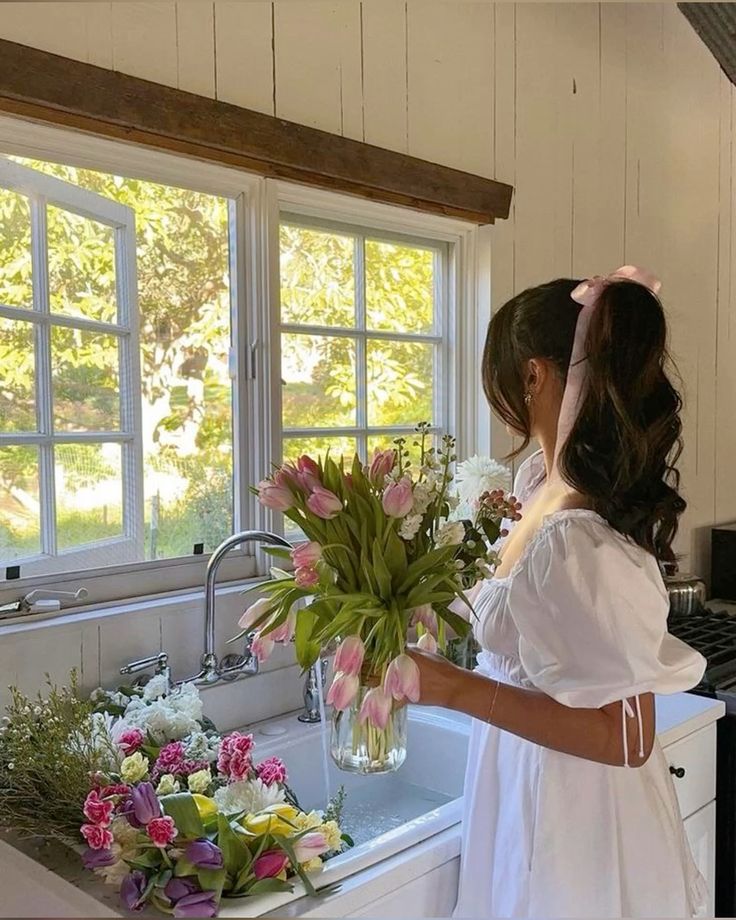 A woman in a white dress holding a bouquet of pink tulips, standing in a bright kitchen by a window with a sink and various flowers nearby.