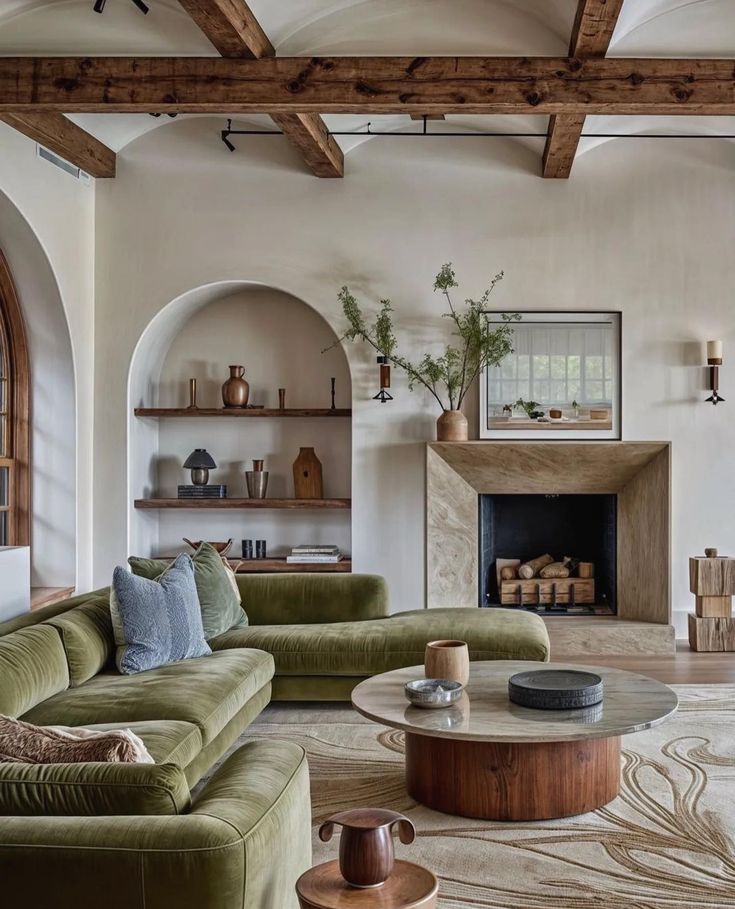 A cozy living room featuring a green velvet sofa, wooden shelves with decorative items, a stone fireplace, and a round coffee table. Natural light enhances the warm, inviting atmosphere.